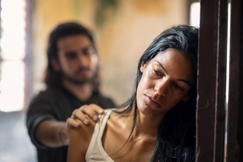 Shoulder and face of a woman with her head resting on a door frame; in the background, a man has his hand resting amiocally on the woman's shoulder.