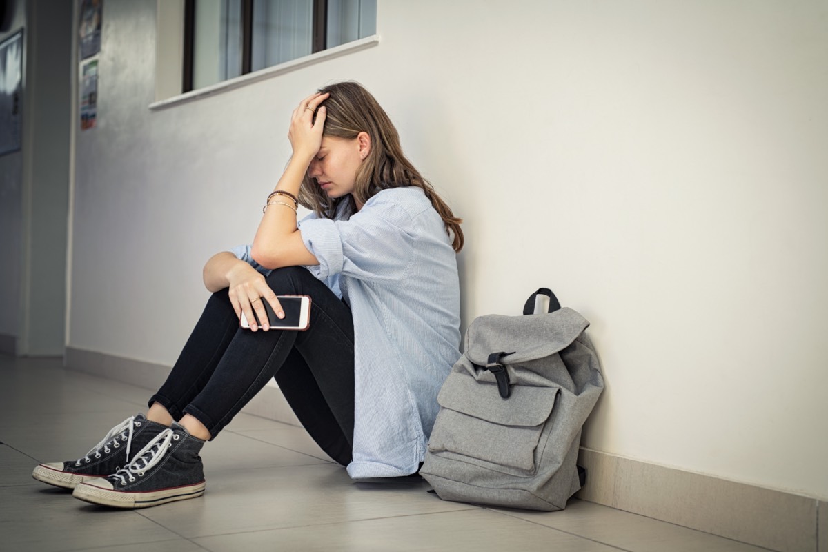 Adolescente déprimée et victime de cyberviolence se tenant la tête avec son smartphone assis sur le sol d'un collège