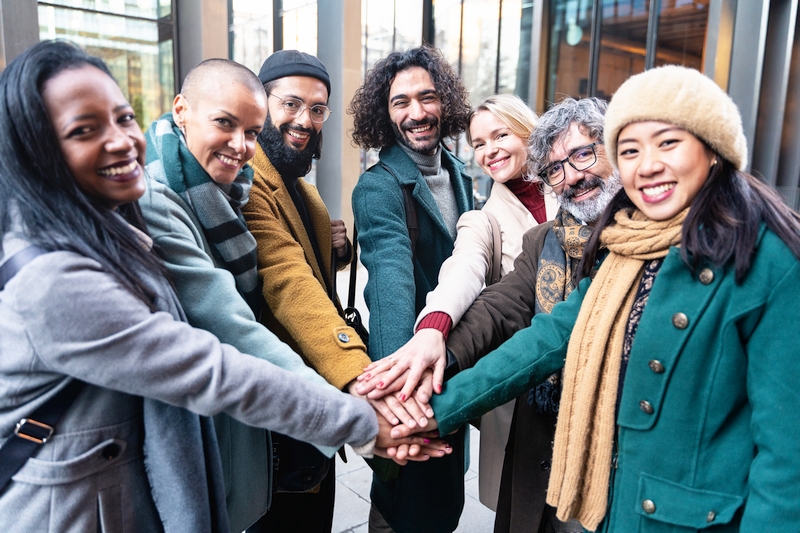 Range of smiling people (dressed in winters), each putting one hand over the other in the middle