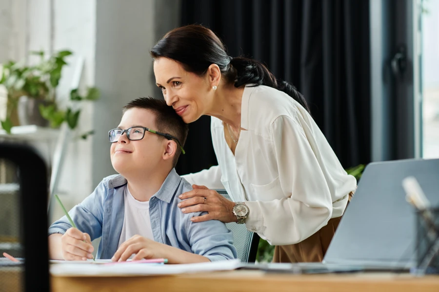 Jeune enfant avec des besoins particuliers travaillant à un bureau sous la supervision bienveillante d’un adulte.